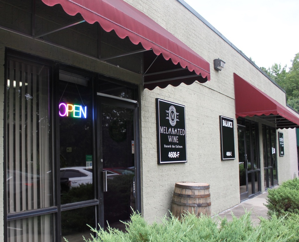 The outside front of Melanated Wine, with a glass door, open sign in the window, maroon awning and a black and white winery sign.