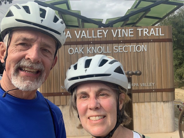 Kyle and Natalie, wearing bike helmets and standing in front of the Napa Valley Vine Trail sign